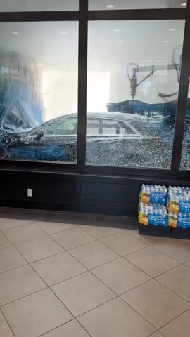 A black sedan is in an automatic car wash. Blue curtains and brushes are visible, along with water spraying onto the car. The car wash appears to be in operation. Outside the car wash, a shelf with snacks and a display of bottled drinks are visible. The floor is tiled.