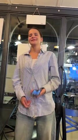A young woman in a blue and white striped button-down shirt and baggy jeans is holding a spoon in her right hand and a phone in her left. She is singing and dancing energetically in front of a glass-paneled wall, possibly a restaurant or cafe. The room is dimly lit with visible light fixtures. She gestures with her arms and body as she sings, occasionally looking up and closing her eyes. In the background, tables, chairs, and some posters are visible through the glass.
