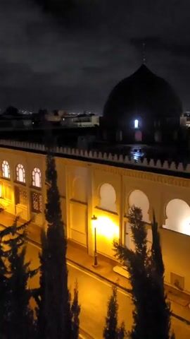 At night, a high-angle static shot reveals a building with a large dark dome atop, its details obscured by darkness, with a single illuminated window. The building's lower section features an arcade of several archways, which are brightly lit from within. A street below is illuminated by lampposts, casting a yellow glow on the wet surface.  Dark trees are visible between the camera and the building, adding a sense of depth. In the background, the city's lights twinkle against a dark, cloudy sky. The building's architecture suggests a Middle Eastern or North African style.