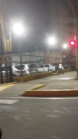 The video shows a nighttime scene of a street. The camera is moving, possibly from inside a vehicle. There are cars parked at what appears to be a toll booth. The camera then passes by what seems to be an outdoor restaurant or food stand with tables and people sitting. A sign saying "Perrotos" is visible on the red structure. There are lights illuminating the area. The ground appears wet, possibly from rain.