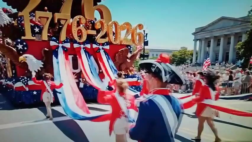 A parade float with the numbers 1776-2026 is depicted, featuring an eagle and patriotic decorations. Several individuals in red and white uniforms are on the float, holding red, white, and blue streamers. In the foreground, people in blue and white uniforms are also holding the streamers. The parade is taking place in an outdoor setting, with a crowd of spectators visible in the background, and a large building with columns on the right side of the frame.