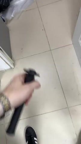 A person's hand is seen holding a black sword. The sword has a black handle and a black blade with a slight curve. The person is wearing a watch on their left wrist. The background appears to be an indoor setting with tiled flooring. The video is short and shows the sword being held steady.