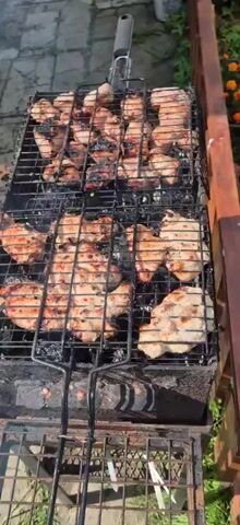 The video shows a close-up, overhead view of chicken pieces grilling on a metal barbecue. The chicken is arranged on two racks of a hinged grill basket, which is placed over a black metal barbecue pit. The chicken pieces are varied in size and shape, with some showing grill marks and others appearing lighter in color, suggesting they are at different stages of cooking. There is visible smoke rising from the barbecue. The background is blurred but appears to be an outdoor setting with greenery and possibly a fence. The lighting suggests it is daytime.