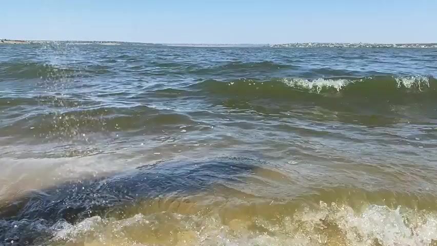 This media file is a video showcasing a serene beach scene in Kyr'yakivka, Ukraine. The primary subjects are the gentle waves of the sea lapping against a sandy shore. The setting is a tranquil beach with a clear blue sky overhead, suggesting a pleasant day.

The activity taking place is the natural ebb and flow of the ocean. Waves are seen forming, cresting with a touch of white foam, and then receding onto the sand, creating ripples and small splashes. The water appears a greenish-brown hue, possibly due to sediment or algae, and the sand beneath the shallow water is a light, warm color with some shells visible.

Notable details include the bright sunlight casting shadows on the seabed and the water, indicating it is daytime, likely midday or early afternoon given the clear sky and bright illumination. The overall mood conveyed is one of peace and natural beauty. There are no people or man-made objects prominently featured, emphasizing the natural environment. The distant horizon shows a faint line of what could be land or a coastline, with a few indistinct structures, providing a sense of place without being intrusive.

No visible text is present in the video. The focus remains on the calming visual of the sea and shore.
