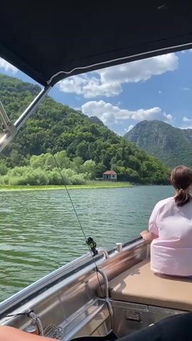 The video opens with a view from the back of a boat on a body of water. A woman in a pink shirt is seated on the right side of the boat, facing away from the camera. The boat has a metal railing and a black canopy overhead. A fishing rod is visible, extending from the boat into the water. The scene shows a lush green hillside covered in trees, with a small, light-colored house with an orange roof nestled among the trees on the shore. The water is green and appears calm, with some lily pads visible. The sky is blue with scattered white clouds. The boat is moving, as indicated by the slight blur and movement of the water and the perspective shift.