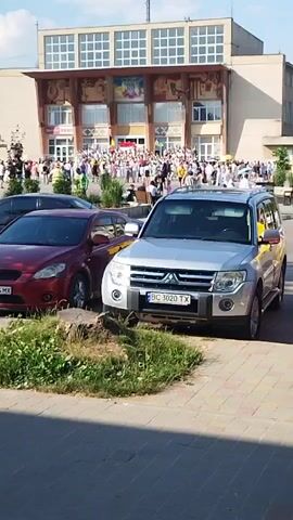 The video shows a crowd of people gathered in front of a building with a large facade and many windows. Several cars are parked in the foreground, including a red sedan and a silver SUV. The sun is shining, and the sky is blue. The people in the crowd appear to be standing and observing something in front of them. Some are holding flags. The cars are parked on a paved area, and there is some greenery and a tree visible. The overall impression is of a public gathering or event.