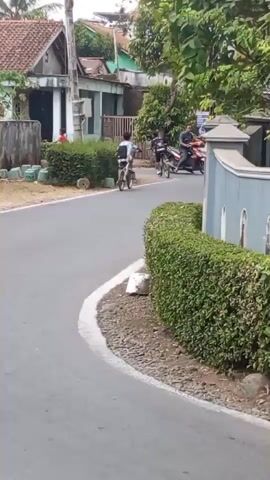 The video shows a street scene. In the foreground, a paved road curves to the right. On the right side of the road, a thick, manicured hedge runs along the curve. On the left side of the road, there is a sidewalk with some greenery and houses in the background. In the middle ground, a person on a bicycle rides away from the camera, heading into the curve. Further in the background, another person on a motorcycle is visible on the right side of the road, also moving into the curve. The environment appears to be a residential area with houses and trees. The lighting suggests daytime.
