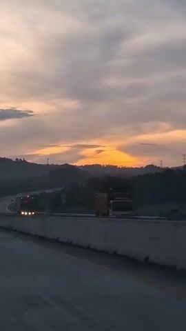 The video shows a highway during sunset. The sky is filled with dramatic clouds in shades of orange, pink, and gray. In the foreground, the highway stretches out, with a concrete barrier separating the lanes. Several trucks and cars can be seen driving on the highway. The landscape on either side of the highway is hilly and covered with trees. The overall mood is peaceful and serene.