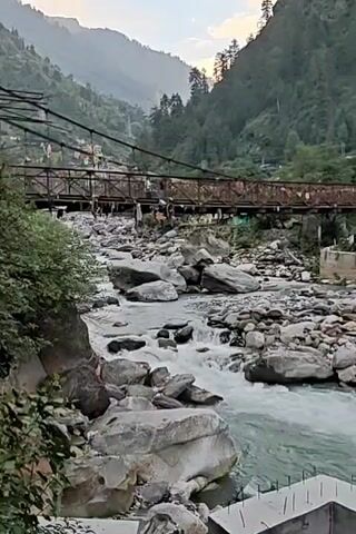 The video presents a serene panorama of a mountain river flowing through a valley. The river, characterized by its light blue-green, turbulent water, is dotted with numerous large, grey rocks. A weathered metal suspension bridge, appearing rusty, spans the river, connecting the two sides of the valley. Several people can be seen walking across the bridge.

On the far bank, colorful multi-story buildings, painted in hues of red, blue, and yellow, are nestled on the hillside, suggesting a vibrant settlement. A prominent cell tower rises behind these buildings. Dense pine or conifer forests cover the surrounding mountains, which rise steeply from the riverbanks into a hazy sky. In the bottom right foreground, a concrete structure with exposed rebar indicates ongoing construction. The soft, diffused lighting suggests it is either late afternoon or early evening. The scene depicts a picturesque blend of natural ruggedness and human activity, located in Manikaran, India. No discernible text is visible.