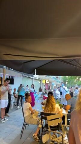 The video shows an outdoor gathering, likely a celebration, with music and drinks. Several people are gathered on a patio-like space with black awnings overhead. Some individuals are seated at tables, while others stand around or mingle. A man with a microphone is speaking or singing, and another man next to him is also holding a microphone. A woman in a long pink skirt stands nearby. There is a bar area decorated with Cuban flags and bunting. People are dressed in casual clothing, and the atmosphere seems lively and relaxed.