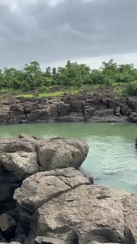 The video displays a river flowing between rocky shores under a cloudy sky. The water is a vibrant green, and the rocks are dark and jagged, forming a natural embankment. Lush green trees and vegetation line the horizon behind the river. The camera pans slowly from right to left, showcasing the rugged landscape. There are no people or animals visible in the frame.