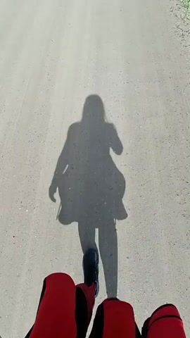 The video begins with a low-angle shot of a shadow of a person on a dirt road. The person appears to be riding a bicycle or motorcycle. The shadow shows the person with their arms raised. As the video progresses, the perspective shifts to show the road ahead, with trees and fields on either side. The sky is blue with some clouds. The shadow of the person's head, holding a phone, is visible on the road ahead. The video captures a sunny day on a rural road.