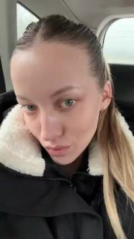 A person with light skin, light hair, and light eyes is sitting in what appears to be a car. They are wearing a dark jacket with a white, fluffy collar. They have a small gold earring in their ear and their hair is pulled back into a low ponytail. The person is looking towards the camera with a slight smile. There is a window visible in the background, showing a blurry scene outside. The overall lighting is slightly dim.