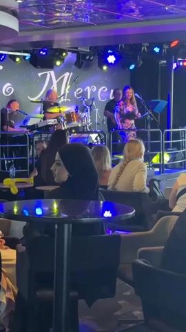 This is a medium shot video of a live music performance in a dimly lit lounge or bar. The stage is set up at the back with a band playing. On the stage, there are musicians, including a drummer and a singer who is interacting with the audience. The band is performing under colorful stage lights, predominantly purple and blue, with some yellow and red accents. The backdrop behind the band appears to have the word "Mer..." lit up in a stylized font. In the foreground, there are several tables and chairs occupied by audience members. The people in the audience are facing the stage, some are seated and observing the performance, while others appear to be engaged in conversation or enjoying drinks. Two cocktails with orange liquid and garnishes are prominently placed on a table in the foreground. The overall atmosphere suggests a relaxed and entertaining evening event.