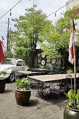 A charming outdoor seating area is depicted. A white Vespa scooter with a sidecar is parked prominently in the foreground on a brick-paved ground. Behind it, several tables and chairs are arranged, some with a vintage sewing machine base. A bamboo pole with a red and white flag, possibly the Indonesian flag, stands near the tables. Lush green trees and potted plants surround the area, providing shade. The background reveals a building with various framed pictures and signs displayed on its exterior wall. The scene appears to be a cafe or restaurant with a retro ambiance, bathed in natural daylight under a partly cloudy sky. No people are visible in the video.