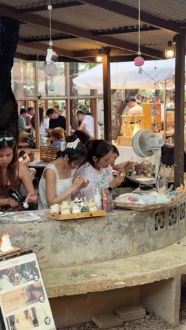 The video shows an indoor-outdoor art studio or cafe setting with people engaged in various activities. In the foreground, two young women are seated at a concrete table, painting small ceramic figures from a tray. The woman on the left, wearing a white lace-trimmed dress, is focused on her work. The woman on the right, in a white t-shirt with green lettering, is also painting and appears to be smiling or interacting with someone off-camera. Several other people are visible in the background, some seated at tables, others standing or walking. The environment is rustic with exposed beams, natural light, and decorative elements like hanging ornaments. A fan is positioned on the table near the painting supplies. A sign with images related to "DIY Modeling" is visible on the lower left.
