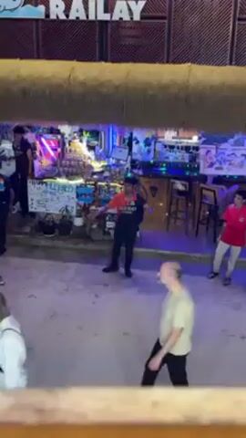 The video shows an outdoor bar called "Railay." Two bartenders stand behind a counter decorated with colorful lights. One bartender is wearing a red shirt and black pants and is gesturing with his hands. Another man in a light green shirt and dark pants is walking in front of the bar and looking towards the camera. The ground is paved with light-colored stones. Above the bar, there is a thatched roof overhang.