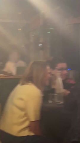 The video shows an interior scene of a dimly lit bar or club with music playing. Several people are present, including individuals sitting at tables and standing near the bar. A DJ can be seen in the background. The bar area is visible with beer taps and bottles. The lighting is low, with some areas appearing hazy or smoky.
