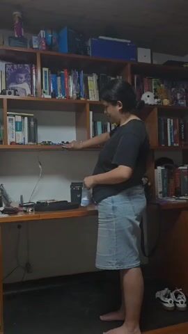 A barefoot, fair-skinned woman in a black t-shirt and light-wash denim skirt stands in front of a wooden desk and bookshelf. She is smiling and touching small toy cars on the shelf. The bookshelf is full of books, small decorations, and framed photos. The desk holds various items including a laptop, cords, and a white paper bag. The room has a low ceiling, and the overall lighting is dim.