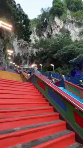 The camera begins its focus at the bottom of a set of brightly painted stairs, then pans up to reveal a gold statue of Murugan, the Hindu God of War, set against a cloudy skyline. The stairs are painted in vibrant shades of red, yellow, blue, and green, with gold accents. The stairs ascend toward a limestone cliff face, adorned with trees and foliage. The lower section of the clip appears to feature artificial lighting in the form of illuminated posts. The area at the bottom of the staircase, behind the statue, appears to be a large open space with many people traversing it.