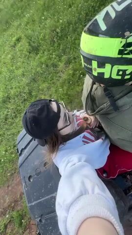Two people are riding on an ATV across a grassy field on a sunny day. The person in the front, holding the camera in a selfie position, is a young woman wearing sunglasses, a baseball cap, and a white sweatshirt. She holds a beer bottle in her hand, smiling and sticking out her tongue at the camera. Behind her, the driver wears a bright green and black helmet and an olive green jacket. The ATV drives along a dirt path through a green grassy area with trees visible in the distance. The sky is blue with some white clouds.