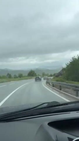 The video shows a view from inside a car driving on a wet road. The windshield wipers are on. There are two cars ahead on the road. There are mountains in the distance and a cloudy sky. There are rocks and green plants on the side of the road. The overall scene is gloomy and rainy.