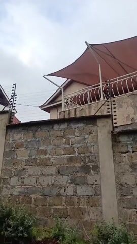 The video shows a residential area. It starts with a close-up shot of a house behind a gray brick wall, focusing on a balcony with a tan sunshade. The camera then pans right to show the house's red tile roof and surrounding fence with security wiring.  The view expands to include more houses behind similar walls along a paved road with a grassy shoulder.  The sky is overcast.  The camera continues moving along the road, keeping the houses and walls in view.