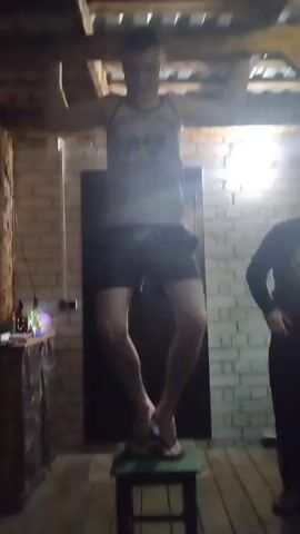 A man stands on a stool, holding onto a wooden beam above him with both hands. He is wearing a tank top and shorts. His legs are bent and his feet are crossed. The stool is green. The background is a brick wall and a wooden floor. There is a door in the background. Another person is partially visible on the right side of the frame. The lighting is dim, with a bright light source visible in the background. The man appears to be struggling to maintain his position.