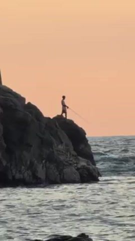 The video shows a person standing on a rocky outcrop by the sea, fishing with a long rod and line. The sky is a muted orange color, suggesting it is either sunrise or sunset. Waves are visible breaking against the rocks and along the shoreline.