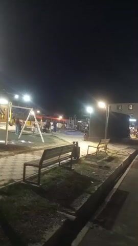 This is a vertical video recorded at night in what appears to be a park or public space. The scene is illuminated by several bright streetlights. In the foreground, there are two park benches facing away from the camera, situated on a patch of grass and dirt. A paved walkway with a cobblestone pattern is visible in front of the benches. In the background, a playground is set up with swings and climbing structures. Several people, including children and adults, are present in the playground area, some interacting with the playground equipment and others standing around. A person is seen on a swing in the center of the frame. To the left, there is a decorative structure with curved arches. The overall atmosphere suggests a public gathering space being used in the evening.