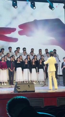 A choir of approximately 25 young individuals performs on a stage indoors. The choir members are dressed in a uniform style: women wear black tops and long white skirts, while men wear black pants and either black tops or white shirts with black bow ties. They are positioned in rows on the stage, singing into several stand-mounted microphones.

A conductor, dressed in a full white suit, stands facing the choir with their back to the camera, actively leading the performance with hand gestures. To the right of the conductor, another individual in a black suit plays a woodwind instrument, possibly a clarinet or oboe.

Behind the choir, a large screen or projection displays an artistic image of two large, silhouetted hands reaching towards each other, with dynamic, flowing colors and patterns. Stage lights are visible above the performance area. Speakers are placed on the stage floor in front of the choir. Part of an audience member's head is visible in the bottom foreground, indicating a live audience. The setting appears to be a formal concert or musical event.