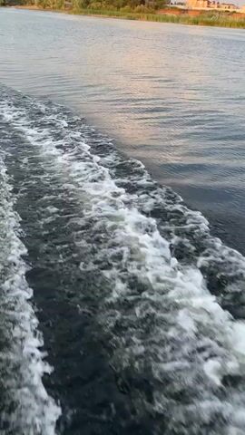 The video shows a close-up view of turbulent water, likely a wake from a boat moving forward. The water is dark with white foam and churned up. The horizon is visible in the background, reflecting the sky, which appears to be a soft, muted color, possibly dawn or dusk. The camera angle suggests the viewer is either on the boat or very close to the water's surface.