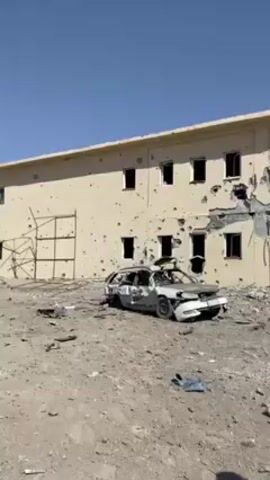 The video shows a war-torn building, featuring numerous bullet holes and structural damage. Debris litters the ground, and a wrecked car sits in the foreground. A man wearing traditional clothing, including a brown robe and a red hat, walks past the camera while talking on a cell phone. The sky is clear and blue, indicating a sunny day.