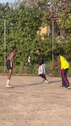 Three men are playing basketball on an outdoor court. The man closest to the camera is wearing a yellow tank top and navy blue athletic pants with red stripes. He dribbles the ball toward the basket. Another man, wearing black shorts and white socks, guards him. A third man in a black shirt and black and white shorts stands further away. The court is made of dirt and gravel. A green, bushy area with tall, white poles forms a backdrop. In the distance is a red roof of a building. The camera follows the man with the yellow tank top as he shoots the basketball toward the basket.