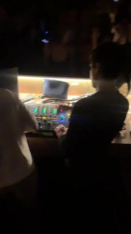 The video opens with a view from behind two individuals operating DJ equipment in a dimly lit club. The DJ booth is illuminated by the equipment's lights, revealing a laptop and various controls. In the background, a crowd of people are visible, some dancing and others socializing. The atmosphere is energetic, with flashing lights and a general sense of excitement. As the video progresses, the camera pans slightly, offering glimpses of the patrons' faces and their interactions. The focus remains on the DJ booth and the lively crowd, capturing the essence of a night out at a club.