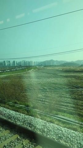 The video, shot from a moving train, captures a rural landscape juxtaposed against a distant cityscape. A railroad track and the gravel beneath it fill the lower portion of the frame. Beyond the track lies a grassy bank, followed by fields. A road with vehicular traffic runs parallel to the train tracks. In the far distance, a cluster of tall buildings forms a cityscape, with hills visible behind them. The sky is a muted turquoise color, and the overall lighting suggests a somewhat hazy day. Bare trees in the foreground offer a glimpse of the scene, moving quickly as the train progresses.