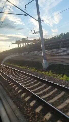 This video shows a view from a moving train. In the foreground, train tracks run diagonally across the screen. The tracks are made of steel rails laid on wooden or concrete sleepers, with ballast beneath. Sunlight casts shadows on the tracks, creating a striped pattern. In the middle ground, there is a green grassy embankment next to the tracks. Beyond the embankment, there is a large pile of what appears to be industrial pipes or similar long, cylindrical objects stacked horizontally. Further in the background, a forest or line of trees is visible. Above the trees, a large industrial structure, possibly a gantry crane, is silhouetted against a blue sky with scattered clouds. Overhead, power lines and utility poles are present, indicating an electrified railway line. The sun is low in the sky, suggesting it is either early morning or late afternoon, and its light creates a lens flare effect in the upper left portion of the video. The camera is positioned to capture the scenery from the perspective of a passenger inside the train.