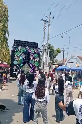 This video shows a street scene with a large sound system on the left, featuring multiple speakers and colorful lights. A group of young people, mostly women, are standing in the street facing the sound system and appear to be dancing or participating in an event. They are dressed casually, with some wearing white shirts with a logo. Tents and stalls are set up along the side of the street, and other people are visible in the background. The weather appears to be sunny.