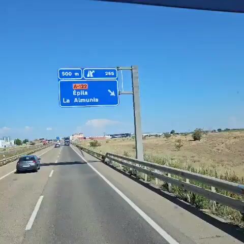 This is a video taken from inside a vehicle traveling on a highway. The video is shaky and appears to be handheld. A blue road sign is visible, indicating "500 m" and "265" to the right, with the road designation "A-122" and destinations "Épila" and "La Almunia." A white car is driving on the highway in front of the vehicle from which the video is taken. The scenery outside the vehicle consists of dry, grassy fields with some scattered bushes and trees. In the distance, there are some buildings, including what appears to be an industrial area or a commercial complex. A speed limit sign indicating "100" is also visible. The sky is clear and blue with a large white cloud formation in the distance. There are no people clearly visible in the video. The overall environment appears to be rural or semi-rural on a sunny day.