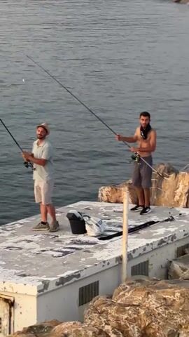 Two individuals are fishing from a concrete structure that juts out into the water, with rocky terrain visible in the foreground and background. The water is calm and appears to be a body of saltwater, likely the sea given the rocky coastline which suggests a coastal or Mediterranean setting. The scene takes place outdoors, under what appears to be natural daylight, possibly early morning or late afternoon given the soft lighting.

The person on the left is wearing a light-colored polo shirt, beige shorts, a straw hat, and casual shoes. They are holding a fishing rod and seem to be in the process of casting or reeling. Next to them is a black bucket and a white bag.

The person on the right is shirtless, wearing grey shorts and black athletic shoes. They also hold a fishing rod, actively engaged in fishing. A dark cloth or scarf is draped around their neck.

The concrete structure they are standing on appears to be a pier or a seawall, with a visible railing or edge. The overall atmosphere suggests a relaxed, recreational activity. There are no visible text elements within the frame. The focus is on the act of fishing and the natural environment.
