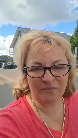 A medium shot captures a woman with blonde hair and glasses from the chest up. She is wearing a pink shirt and a gold chain necklace. Her expression is one of displeasure. The background shows houses and trees under a cloudy sky.