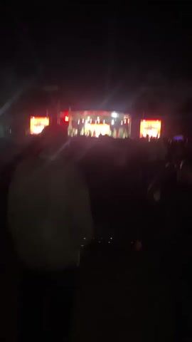 The video shows a concert at night. A large crowd of people faces a brightly lit stage. Several people are visible on the stage, likely performers. There are two large video screens on either side of the stage, displaying images. Bright lights shine down from above the stage. A person is standing in the foreground with their back to the camera. The overall environment suggests an outdoor music festival or concert.