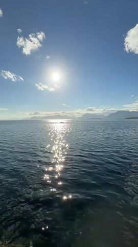 A wide shot shows a body of water under a bright sun. The sun is high in the sky, casting a shimmering path across the water. In the distance, mountains are visible on the horizon. The water is a deep blue, with some lighter green hues near the shore. The sky is clear and blue, with a few fluffy white clouds. There is a small boat with two people visible in the distance. The video appears to have been shot from a low angle, looking out over the water. The overall impression is one of a peaceful and serene day.