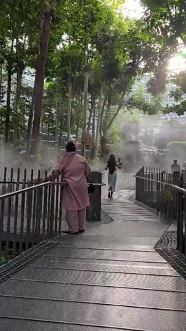 The video begins on a metal walkway with railings, looking towards a lush green area with trees and a man-made waterfall emitting mist. In the foreground, a person wearing a pink outfit stands facing away from the camera, leaning on the railing. Further down the walkway, a person in a blue top and white pants walks away from the camera. In the background, several other people are visible, some sitting and others standing near the waterfall. The sun is shining through the trees, creating a bright effect. The walkway appears to be part of an outdoor or semi-outdoor park or garden space.