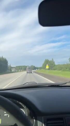 The video captures a first-person perspective from inside a car driving on a multi-lane road in Marchugi, Russia. The dashboard, steering wheel, and a rearview mirror are visible in the foreground, with the windshield framing the view ahead. The car is traveling on a clear, paved road, marked with solid and dashed white lines.

Ahead on the road, a dark-colored van is visible, driving in the same direction. To the left, a Lukoil gas station can be seen in the distance, featuring its red and white branding and fuel pumps. Prices are partially visible on its sign. The roadside is lined with green grass and numerous trees, suggesting a rural or semi-rural environment.

On the right side of the road, a yellow triangular warning sign with a red border and a black "A" symbol (indicating a crossroad or intersection) is prominent. Another similar yellow triangular sign is partially obscured behind it. The sky is bright with scattered white clouds.

Inside the car, the speedometer is visible on the dashboard, showing increments up to 200 km/h, with the needle indicating a speed of approximately 80 km/h. A trip meter displays "297.4" and "333". The sound of music is playing in the background throughout the video.