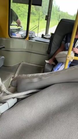 The video shows the interior of a bus from the perspective of a passenger, looking towards the driver's seat and the front of the bus. In the driver's seat, a person wearing a dark t-shirt is visible in profile, with their hands on the steering wheel. To the right of the driver's seat, an older woman is seated, holding a white pillow and a small white object, possibly a card or ticket, in her hands. She is wearing a striped shirt. The bus appears to be in motion, with a view of trees and a road through the windshield. The interior of the bus is utilitarian, with gray seats and yellow accents on the dashboard and some interior panels. The foreground is dominated by a textured gray fabric, possibly a seat cover or a bag.