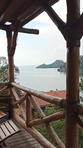 From a balcony constructed of bamboo and rope, a view of a body of water, possibly a lake, is visible. A small island covered in trees sits in the middle distance. The sky is overcast. Below the balcony is a red-tiled roof and lush greenery. There are several wooden chairs on the balcony.