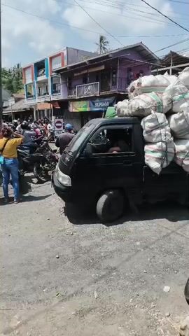 A black pickup truck is parked on a dusty road. The truck is loaded high with many large, white sacks tied with string and green and red bands. A person is visible in the driver's seat. To the left of the truck, several people are standing near motorcycles and a market stall with colorful awnings. The background shows buildings, power lines, and trees under a bright sky.