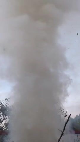 The video shows a thick plume of gray smoke rising into the sky against a light gray, overcast sky. The smoke is dense and appears to be ascending rapidly, creating a funnel-like shape at its base. Small dark particles, possibly debris, are visible scattered within and around the smoke.

The scene is set outdoors, with the tops of trees and branches visible at the bottom of the frame. The foliage suggests it might be late spring or summer. In the lower right corner, a thin, dry branch points upwards, partially obscuring the smoke. There are also hints of structures or rooftops in the far background, particularly on the right side.

The overall impression is of a significant combustion event, possibly a fire or an explosion, that has generated a large amount of smoke. The time of day appears to be either early morning or late evening, given the muted light and the color of the sky. There is no visible text in the media. The emotion conveyed is one of potential danger or a dramatic event unfolding. The location context is Великово, Russia.