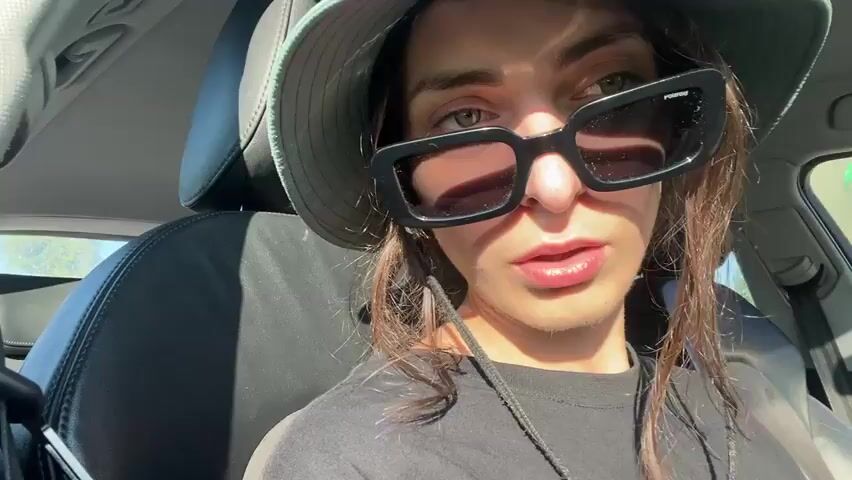 A close-up shot captures a person wearing a green bucket hat and black sunglasses, sitting in the driver's seat of a car. The person looks directly at the camera with a slight smile, their hair falling around their face. Sunlight casts shadows through the sunglasses, obscuring their eyes. The background shows the interior of the car and a blurred view of a residential area through the windows.